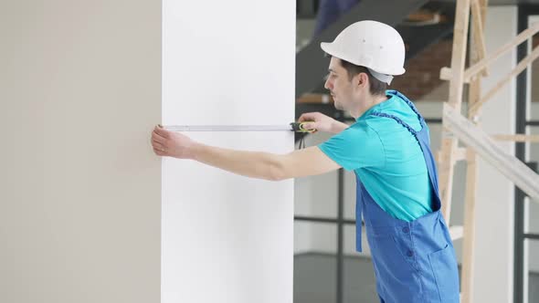 Side View of Professional Concentrated Caucasian Man in Hard Hat and Uniform Measuring Wall Indoors