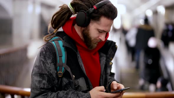 Young Handsome Man with Dreadlocks is Listening to Music By Headphones and Phone in Metro Station