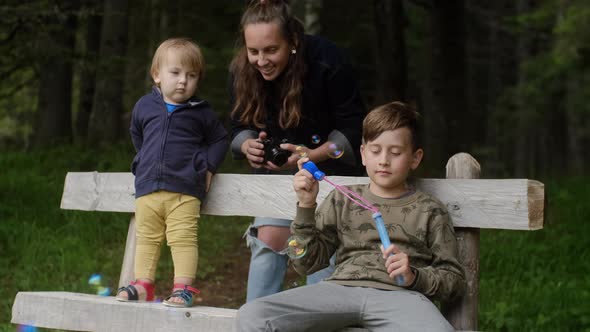 Young mother with her sons playing with soap bubbles and makes pictures on the camera in the park.