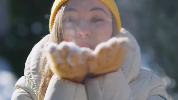 Closeup Smiling Face of Young Charming Woman Blowing Snow From Yellow Mittens