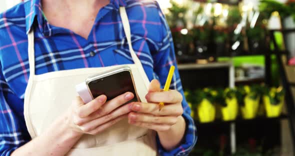 Female florist writing an order on notepad while using mobile phone