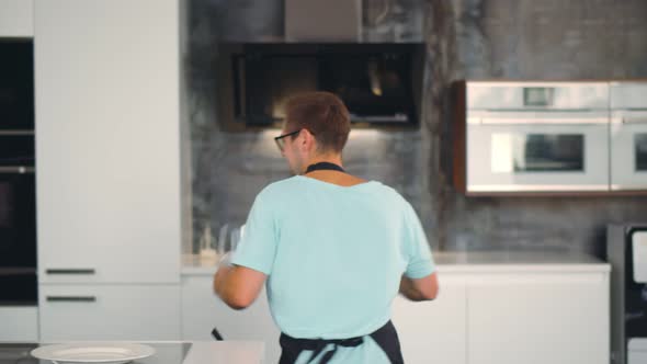 Young Handsome Man in Apron Dancing with Wine Glasses Setting Table for Romantic Dinner