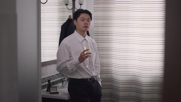 Unsure Asian Groom Standing in Bathroom with Glass of Champagne Thinking