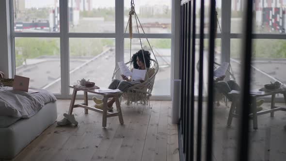 Extreme Wide Shot of African American Woman Sitting on Hanging Chair Analyzing Paperwork