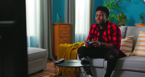 Black AfricanAmerican Excited Man Sitting on a Sofa Holding a Joystick Playing Video Games