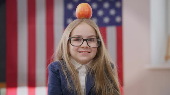 Charming Joyful Teen Schoolgirl Looking at Camera Smiling with Apple on Head and USA Flag at