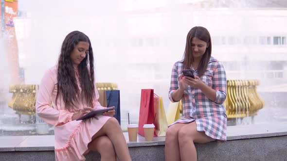 Two Girls After Shopping are Sitting in the Park Near the Fountain Using a Phone and a Tablet Having