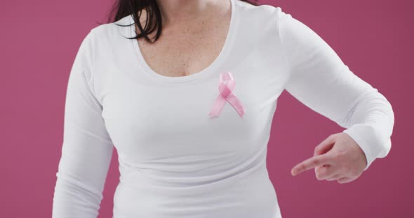 Mid section of a woman pointing to the pink ribbon on her chest against pink background