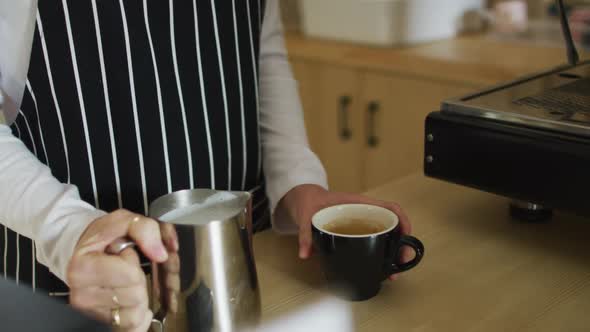 Midsection of caucasian waitress wearing apron, pouring milk into cup of coffee