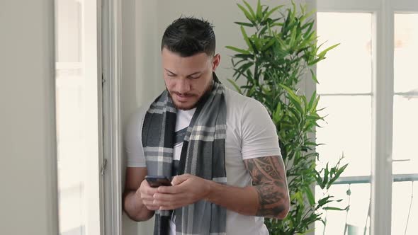 Thoughtful Bearded Young Man Standing with Smartphone Near Window.