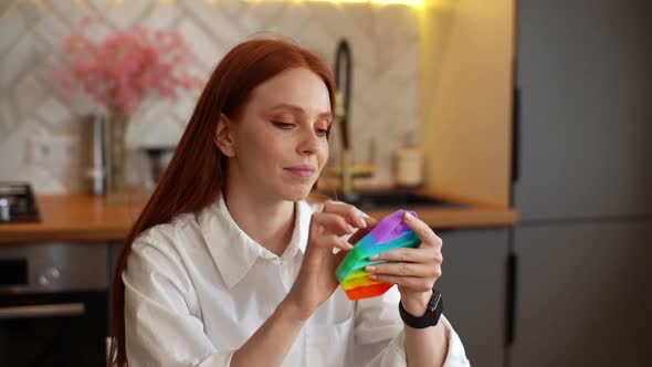 Portrait of Cheerful Redhead Young Woman Playing with Rainbow Popit Fidget Toy Sitting at Table in