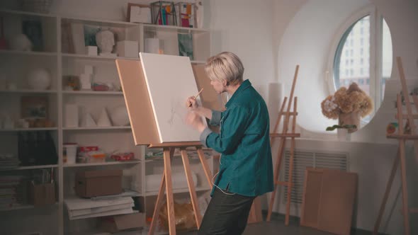 A Young Woman with Short Blonde Hair Drawing Sketch on the Canvas and Listen Music in the Headphones