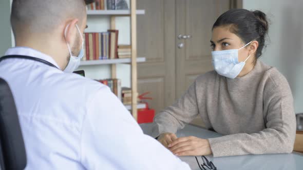 Male doctor in protective mask and lab coat sitting at desk in clinic, reading medical history