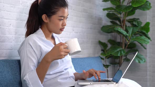 Young Asian Woman Working on Laptop While Holding and Drinking Coffee Indoors