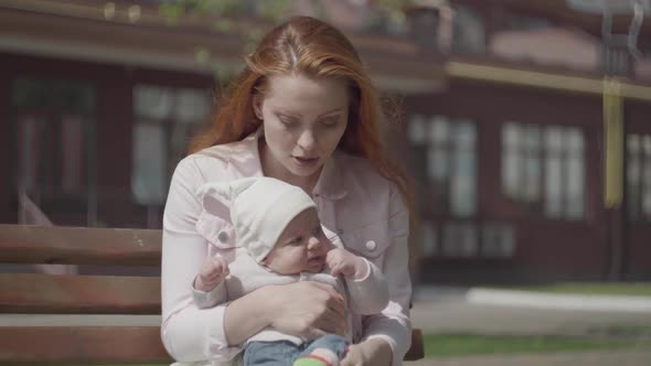 Portrait of a Beautiful Red-haired Woman Playing with Her Child Sitting on the Bench Close-up. The