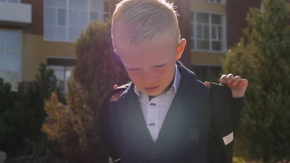 A Little Boy in a Shirt and Jacket with a School Bag Near the School Building