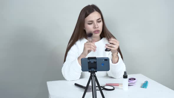 A Caucasian Girl Blogger in Front of a Cellphone Puts a Blush on Her Face Brush.