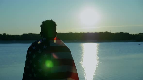 Afroamerican Man with American Flag on Shoulders Looks Into Distance at Sunrise