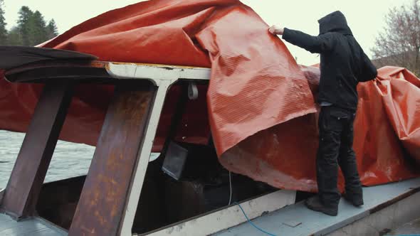 Young man untying  waterproof cover of wooden boat before starting restoration maintenance