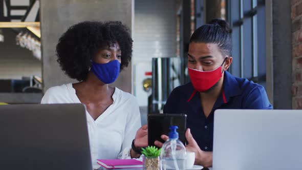 Diverse couple wearing face masks sitting in cafe using laptops and smartphone