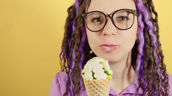Young woman in glasses eating ice cream on yellow background.
