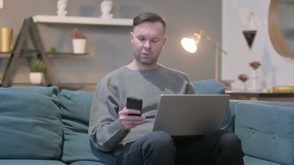 Casual Man with Laptop Using Smartphone on Sofa