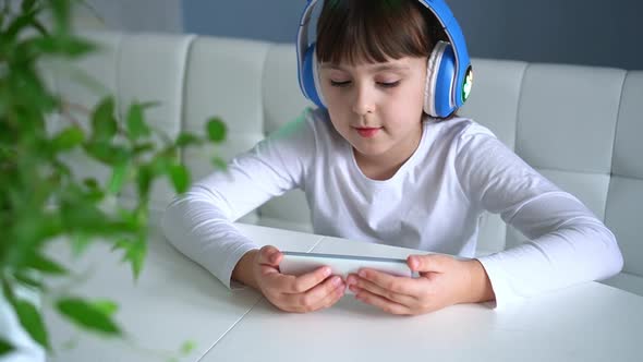 Close Up of Child in Blue Headphones Listening to Music on White Smartphone on Sofa in Living Room