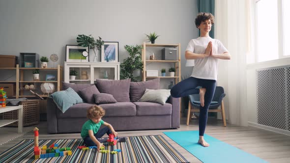 Young Woman Practising Yoga at Home While Calm Child Playing with Toys on Floor