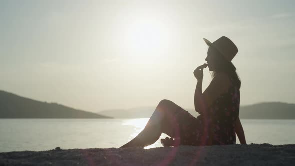 A Girl in a Dress and a Hat Eats Strawberries at Sunset By the Sea