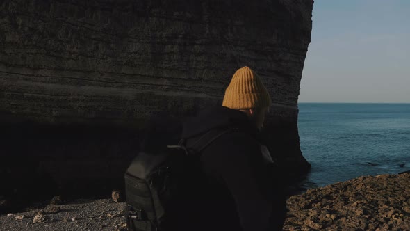 Happy Young Male Tourist with Backpack Walks Along Sunny Rocky Beach at Etretat Towards Famous White