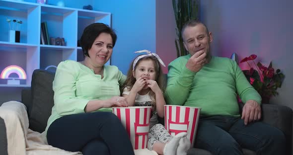 Senior Caucasian Сouple Sitting on Sofa at Home with 5 Year Old Granddaughter and Eating Popcorn