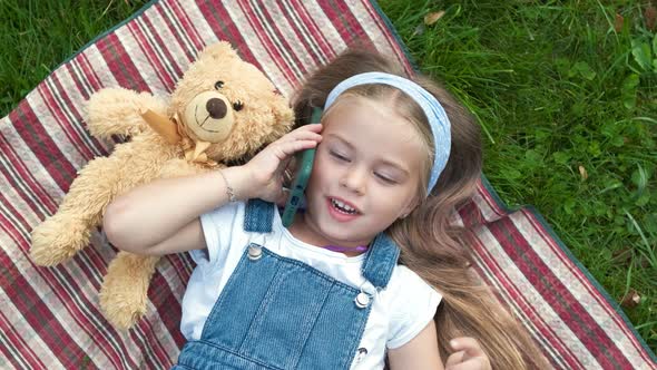 Little Happy Child Girl Laying on Green Lawn with Her Teddy Bear Talking on Mobile Phone