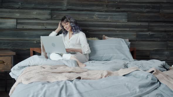 Slow Motion of Cheerful Young Woman Using Laptop in Bed and Smiling Enjoying Internet Content