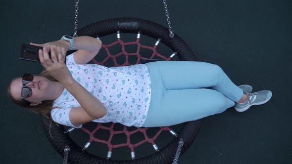 a Young Married Girl in Sunglasses Lies on a Homemade Swing and Writes a Message on the Phone