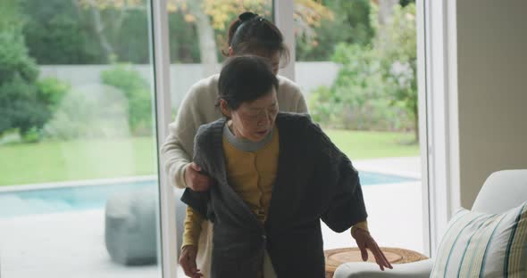 Senior asian mother being helped to armchair by adult daughter in living room