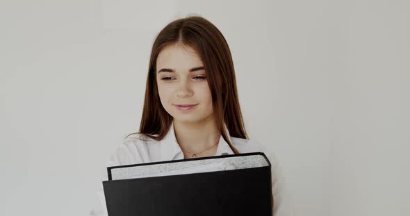 Portrait of Girl with Folder in Hands Looks with Smiles at Camera on Background