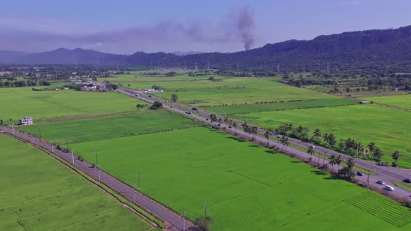 Aerial view of driving cars on road beside Agricultural Fields and smoking stacks behind the mountai