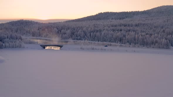 Aerial view tracking vehicle driving across misty ice lake bridge in snow covered Scandinavian woodl