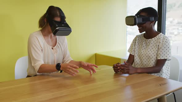 Two diverse female coworkers sitting at desk, testing vr googles