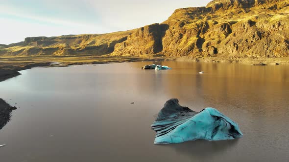 Drone Over Lagoon With Blue Ice Towards Sunlit Rocky Landscape