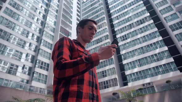 Young Man Uses a Smartphone on a Background of a Residential Skyscraper Highrise Building