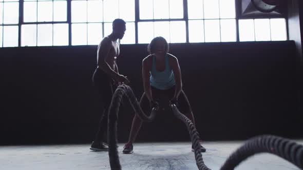 African american woman exercising battling ropes in empty urban building with man cheering her on