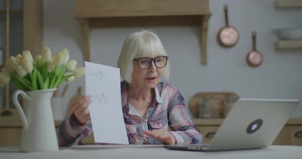 Senior Female Teacher in Glasses Talks and Shows Sheet of Paper with Physics Equations Looking at