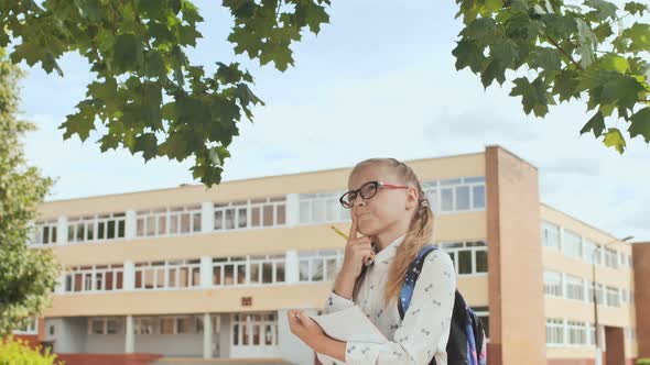 Little Schoolgirl Writes in a Notebook Against the Background of Her School.