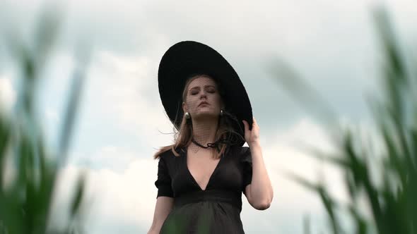 Blonde Holds Big Black Hat With Her Hand On Her Head.