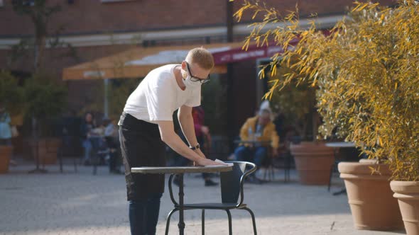 Waiter Wearing Protective Face Mask Disinfecting Table at Outdoor Cafe