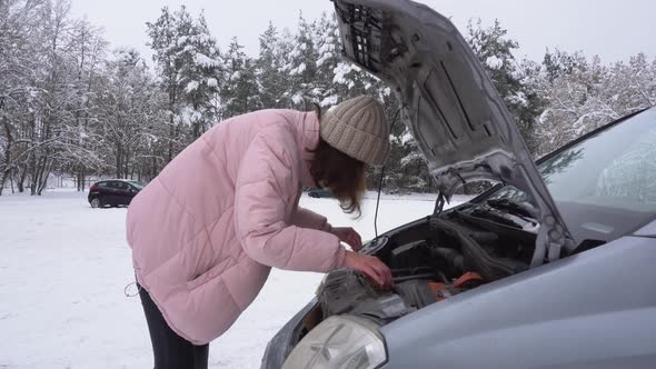 A Girl in a Pink Winter Jacket and a White Hat Inspects a Broken Car Under the Hood in Winter