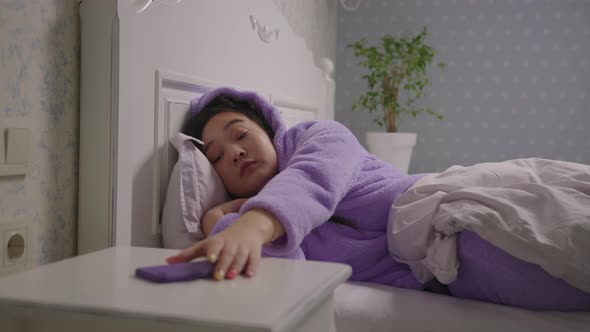 Asian Woman in Purple Pajamas Waking Up and Taking Mobile Phone in Hands Lying in Bed