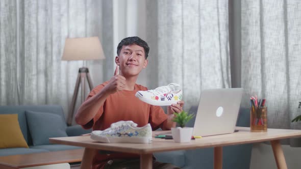 Smiling Asian Boy Footwear Designer Holding Sneaker And Showing Thumbs Up Gesture To The Camera