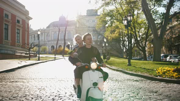 Happy Young Hipster Couple Riding a Vintage Scooter in Old City Center with Grocery Paper Bag